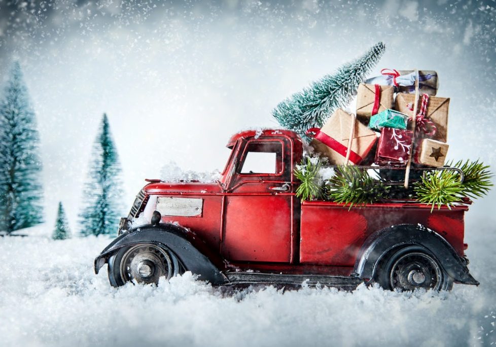 Festive red vintage truck laden with Christmas gifts and pine tree for decorating driving through a winter snow storm in a greeting card design with copy space
